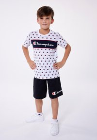 White cotton t-shirt with red and navy Champion logo, paired with black shorts featuring logo; child stands confidently, hands on hips.
