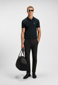 Svart polo skjorta med ett diskret mönster, korta ärmar och krage; kombinerad med svarta slim-fit byxor och en svart duffelväska.