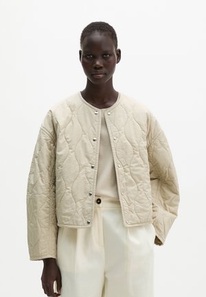 Persoon draagt een beige doorgestikte jas met knopen over een licht topje en crèmekleurige broek met hoge taille, tegen een effen witte achtergrond.