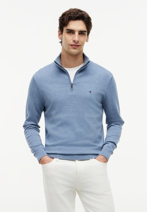 Mladý muž nosiaci svetlomodrý sveter so zipsom po polovicu a bielu košeľu s bielymi nohavicami, ruky v vreckách, na jednoduchom pozadí.