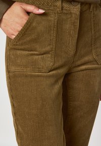 Pantaloni in corduroy marrone con righe verticali, tasche laterali e vita sagomata. Il tessuto sembra morbido con una finitura testurizzata.