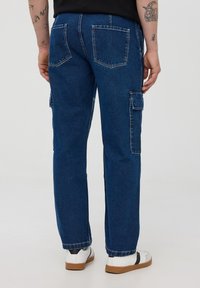 Spodnie cargo z denimu w ciemnoniebieskim kolorze, o prostym kroju nogawki, z widocznymi tylnymi kieszeniami i kieszeniami cargo po bokach. Charakteryzują się minimalnym przeszyciem i gładką teksturą.