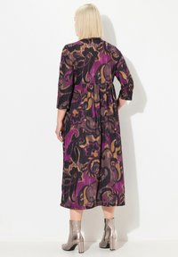 Robe maxi à motifs violets et noirs avec des manches trois-quarts ; elle présente une taille froncée et une silhouette fluide, associée à des bottines métalliques.