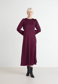 Burgundy satiini pitkähihaisessa mekossa on tiukka vartalo-osa ja virtaava hame, yhdistettynä mustiin teräväkärkisiin saappaisiin. Liukas tekstuuri, minimaalinen muotoilu.