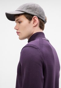 Μωβ πουλόβερ με ψηλό γιακά και ραφές στους ώμους, συνδυασμένο με γκρίζο καπέλο με καμπυλωτό γείσο και λογότυπο στο πίσω μέρος.