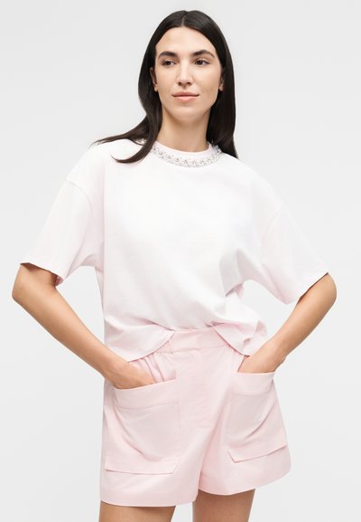 Γυναίκα με μακριά σκούρα μαλλιά φοράει μια φαρδιά λευκή μπλούζα με διακοσμητικό λαιμόκοψη και ανοιχτό ροζ σορτς με μεγάλες μπροστινές τσέπες, χέρια στις τσέπες.