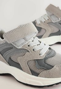 Lekkie buty sportowe z szarym zamszem i siateczką, białymi sznurowadłami oraz trwałą czarną gumową podeszwą. Rzep dla zapewnienia pewnego dopasowania.