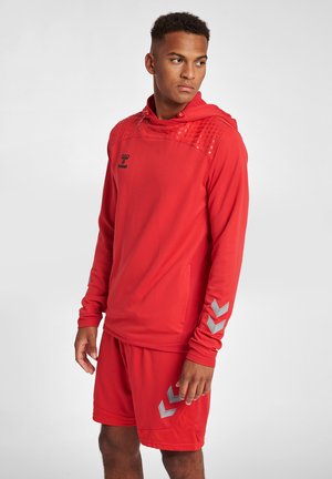 Ung mand i rødt sportstøj med hættetrøje med lange ærmer og matchende shorts, stående og kigger til siden mod en ensfarvet baggrund.