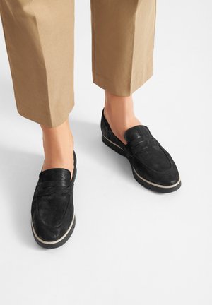 Melni zamšādas mokasīni ar teksturētu virsmu, izrotāti ar šuvju dizainu un kontrastējošu gaišāku zoli. Valkāti kopā ar brūniem, nedaudz saīsinātiem biksēm.