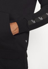 Felpa nera in misto cotone, con tasca frontale, maniche con polsini e dettagli decorativi con loghi Puma lungo il braccio.