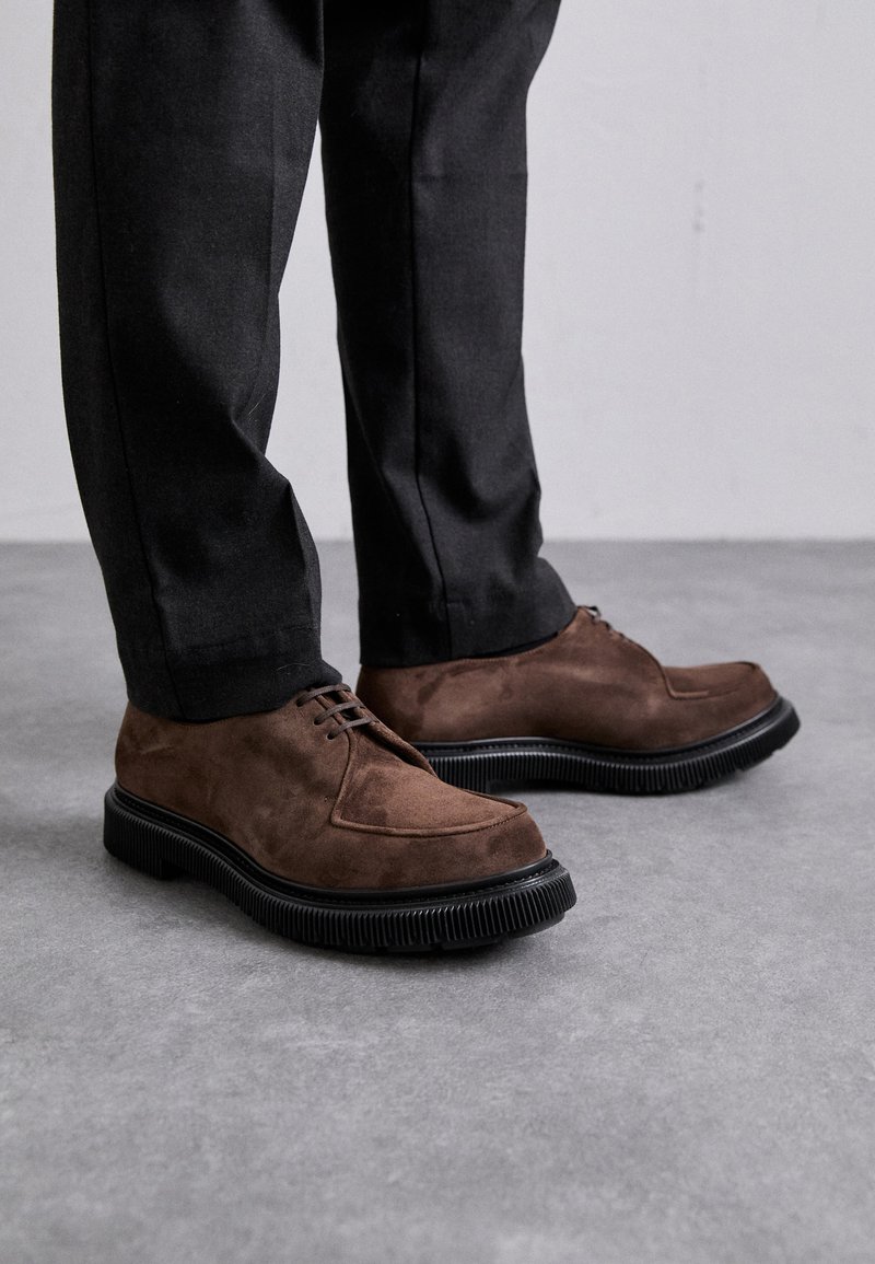 Brown suede shoes with a rounded toe, laced design, and thick black rubber sole. Worn with black tailored trousers against a gray background.