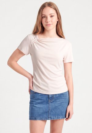 Jeune femme aux longs cheveux châtain clair portant un haut rose clair à manches courtes et une jupe en jean bleue, debout avec une main sur la hanche.