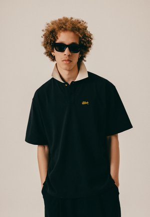 Jauns cilvēks ar cirtainiem matiem, valkājot melnas saulesbrilles un melnu polo kreklu ar bēša apkakli, stāv ar rokām kabatās.