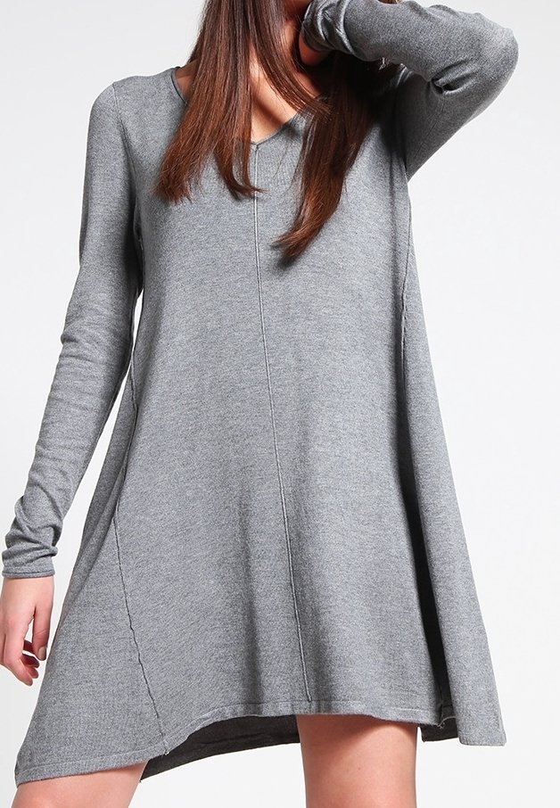 Femme portant une robe grise ample à manches longues avec un col en V, debout avec un bras levé vers sa tête.