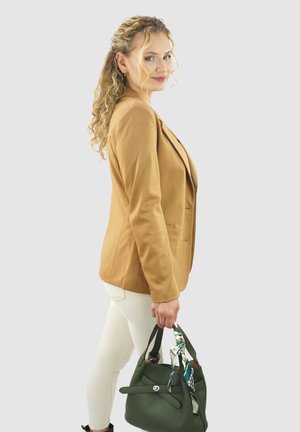  Lichtbruine blazer met een getailleerde pasvorm, gecombineerd met witte broek. Een groene handtas heeft een decoratieve sjaal die aan het hengsel is geknoopt.