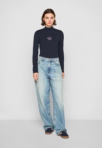 Marineblauwe langemouwen turtleneck met wit logo, gecombineerd met hoog in de taille gesneden lichtblauwe wijde jeans. Model draagt marineblauwe sneakers.