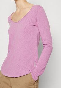 Pitkähiha pinkki paita, jossa on V-pääntie, valmistettu pehmeästä, teksturoidusta kankaasta. Sisältää sivusauman tarran ja rento leikkaus.