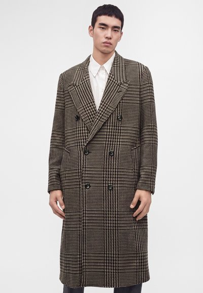 Jeune homme portant un long manteau à carreaux marron et noir à double boutonnage par-dessus une chemise blanche à col, debout devant un fond clair uni.
