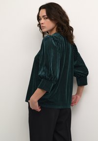 Blusa di velluto teal con strisce verticali, maniche a sbuffo e vestibilità morbida. Il tessuto ha una texture lucida, che enfatizza il suo colore ricco.