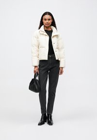 Bijela puffer jakna, crni top, crne traperice visokog struka i crne gležnjače. Model drži malu crnu torbicu. Jednostavan dizajn, strukturirani kroj.