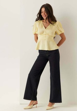 Donna che indossa una blusa gialla con maniche a sbuffo, jeans scuri a gamba larga e scarpe beige con tacco e cinturino dietro, in piedi con le mani dietro la schiena.