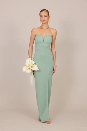 Kvinne i en skulderløs salviegrønn kjole som holder hvite kalla-liljer, iført gulløreringer og armbånd, stående foran en beige bakgrunn.