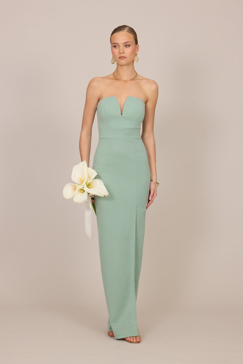 Γυναίκα με φόρεμα σε πράσινη απόχρωση φασκόμηλου χωρίς τιράντες, κρατώντας λευκά κάλλα, φορώντας χρυσά σκουλαρίκια και βραχιόλι, στέκεται μπροστά σε μπεζ φόντο.