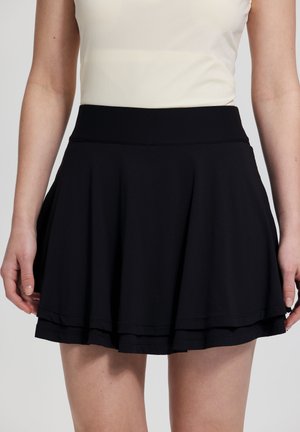 Kvinde iført sort lagdelt skaternederdel og en hvid ærmeløs top, vist fra taljen til midt på låret mod en ensfarvet baggrund.