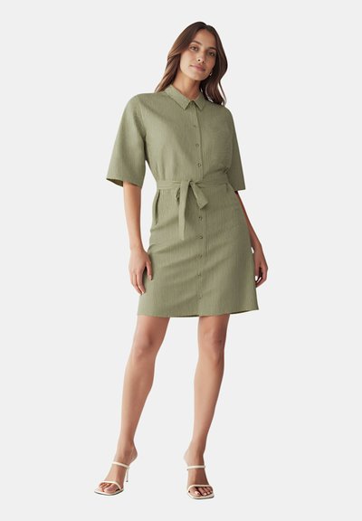 Abito verde con maniche corte, apertura con bottoni e vita annodata. Tessuto testurizzato e taglio al ginocchio. Abbinato a sandali con tacco bianchi.