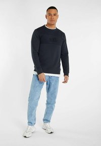 Sweat-shirt marine avec logo NXG texturé sur la poitrine, col rond et poignets côtelés. Associé à un jean en denim bleu clair et des baskets blanches.