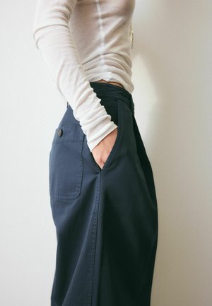 Person wearing a white long-sleeve top and dark high-waisted pants with a buttoned back pocket, hand in side pocket, against a plain background.