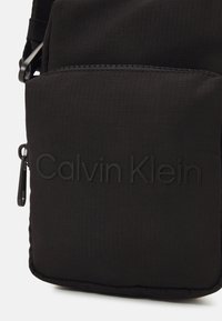 Zaino nero con un tessuto testurizzato che presenta un grande logo "Calvin Klein" in rilievo. Include una tasca con zip per ulteriore spazio di archiviazione.