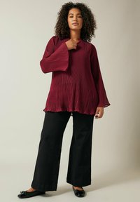 Blouse plissée bordeaux avec des manches évasées et un col haut, associée à un pantalon noir à jambes larges et des chaussures plates noires. Texture de tissu lisse.