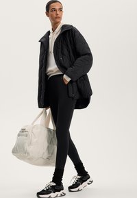 Steppende zwarte jas, roomkleurige hoodie, zwarte legging en zwarte sneakers met witte accenten. Lichtgekleurde duffeltas in de hand gehouden.