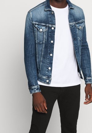 Uomo che indossa una giacca di jeans blu sopra una maglietta bianca e pantaloni neri, in piedi davanti a uno sfondo chiaro e uniforme.