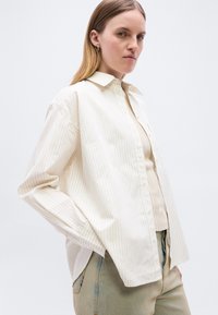 Γυναίκα με ίσια ξανθά μαλλιά, φορούν μια άνετη λευκή πουκαμίσα με λεπτές ρίγες πάνω από μπεζ τοπ και ανοιχτόχρωμο παντελόνι με κεντήματα.