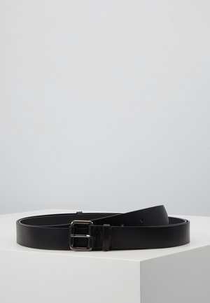 Musta nahkavyö hopeanvärisellä suorakulmaisella soljella, kierretty valkoisen jalustan päällä vaaleanharmaata taustaa vasten.