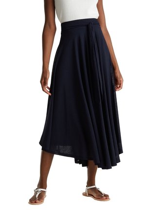 Femme portant une jupe midi asymétrique bleu marine nouée à la taille, associée à un haut blanc sans manches et des sandales blanches plates.