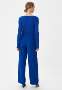 Kobieta z włosami związanymi w kok, ubrana w dopasowany niebieski sweter prążkowany, wysokie, szerokie niebieskie spodnie i niebieskie sandały na obcasie, odwrócona tyłem.