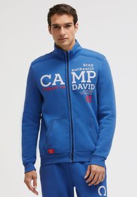 Blue zip-up sweatshirt made of soft material, featuring white and red text graphics, ribbed hem, and two front pockets.