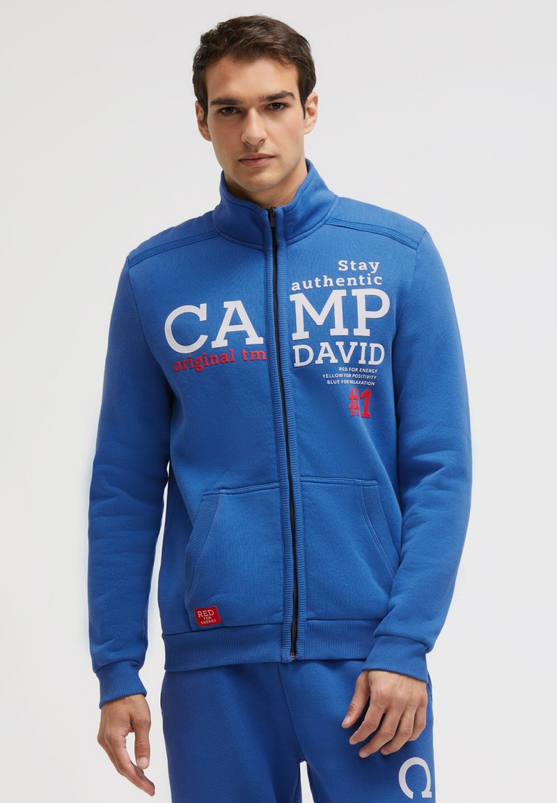 Blue zip-up sweatshirt made of soft material, featuring white and red text graphics, ribbed hem, and two front pockets.