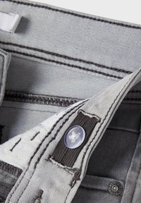 Light grey denim fabric with dark grey stitching, featuring a button closure, and a zipper. Smooth texture, showing interior pocket and label.
