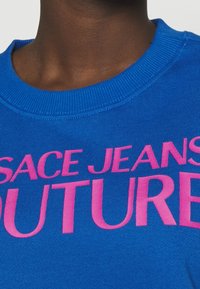 Gros plan sur un sweat-shirt à col rond bleu côtelé avec un texte rose vif indiquant "SACE JEANS FUTURE."