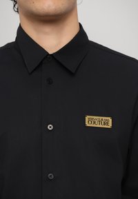 Black button-up shirt with a structured collar, featuring a gold logo tag that reads "VERSACE JEANS COUTURE." Smooth texture, classic fit.
