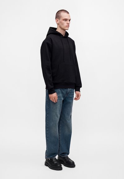 Jeune homme debout portant un sweat à capuche noir, un jean bleu coupe décontractée et des chaussures noires épaisses sur fond blanc uni.