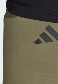 Zelené legíny s texturovaným žebrovaným materiálem, přiléhavého střihu, s černým logem Adidas na boku a černým pasem nahoře.