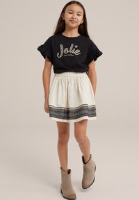 Musta lyhythihainen paita, jossa on frillireunat ja kultainen "Jolie" -teksti, yhdistettynä kermaiseen hameeseen, jossa on musta kuvioreuna ja beigeet saappaat.