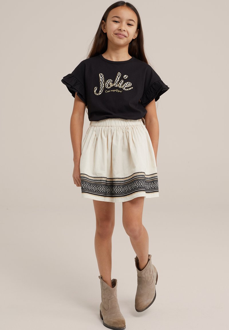 Musta lyhythihainen paita, jossa on frillireunat ja kultainen "Jolie" -teksti, yhdistettynä kermaiseen hameeseen, jossa on musta kuvioreuna ja beigeet saappaat.