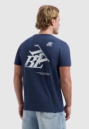 Jonge man met blond haar staat met de rug naar de camera, draagt een marineblauw T-shirt met witte grafische tekst en lichtblauwe spijkerbroek.