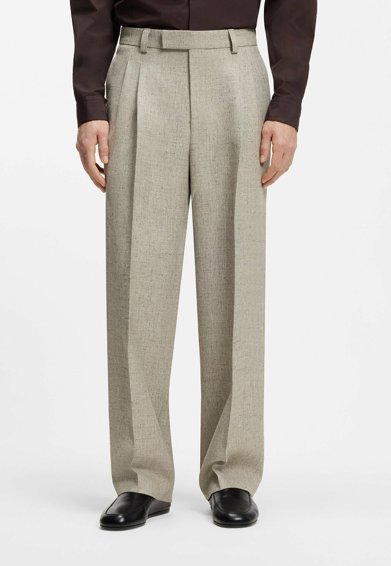 Homme portant un pantalon beige taille haute à jambes larges avec des plis sur le devant, associé à des chaussures en cuir noires et une chemise sombre à manches longues.